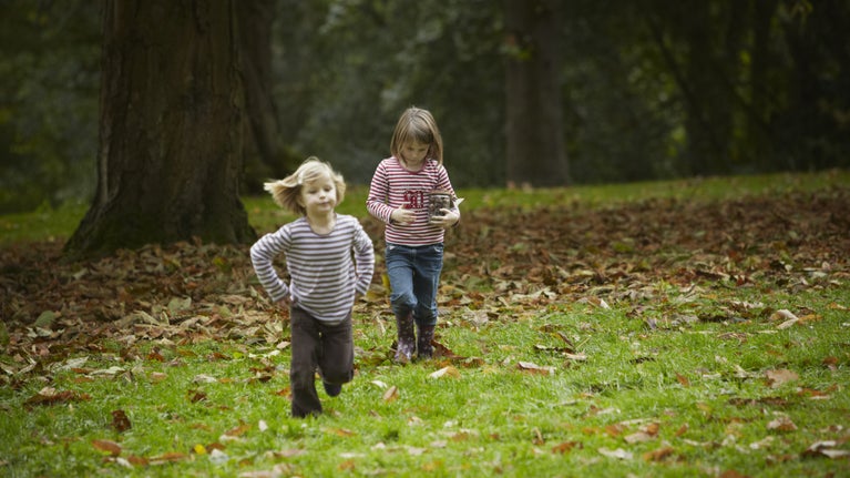 Two children walk towards the camera, over a lawn covered in fallen leaves.